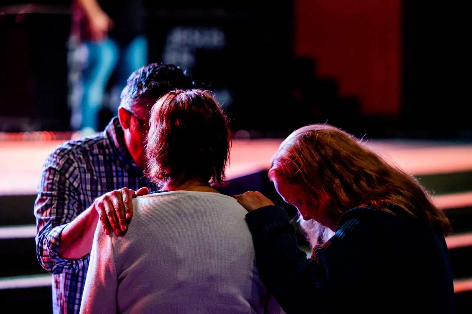 Three adults with hands on one another’s shoulders during prayer in a church